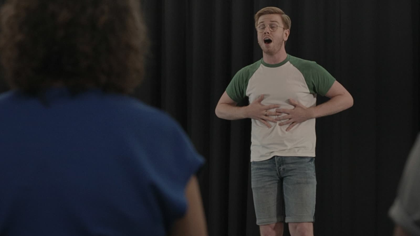 A performer mid-song in the Secret Sing room, hands on his ribs, the group watching from the front.